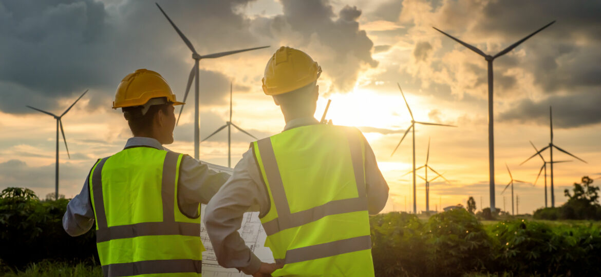 rear view of two asian engineers wearing safety vest holding blueprint near wind turbine field during sunset